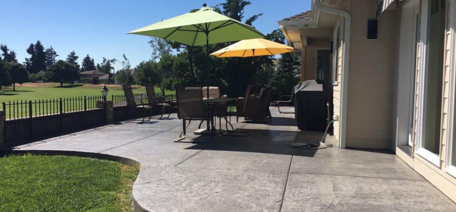 Stamped concrete patio with outdoor dining furniture and umbrellas beside a home overlooking a landscaped backyard.