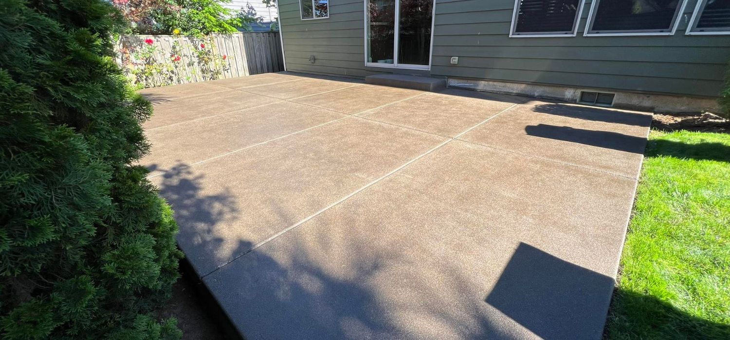 Newly installed broom-finish concrete patio behind a residential home with clean control joints, bordered by grass and landscaping in a backyard setting.